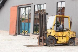 Old yellow, black, rusty scratched forklift truck parked on road near a big industrial warehouse.